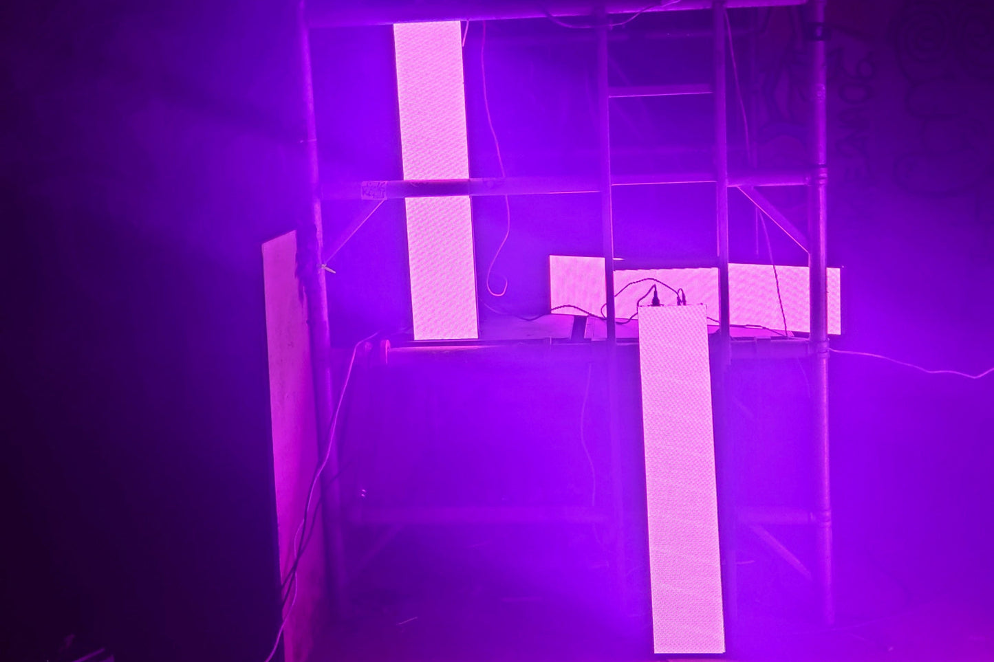 3 LED panels glowing pink on scaffolding in a warehouse with power cables in the foreground
