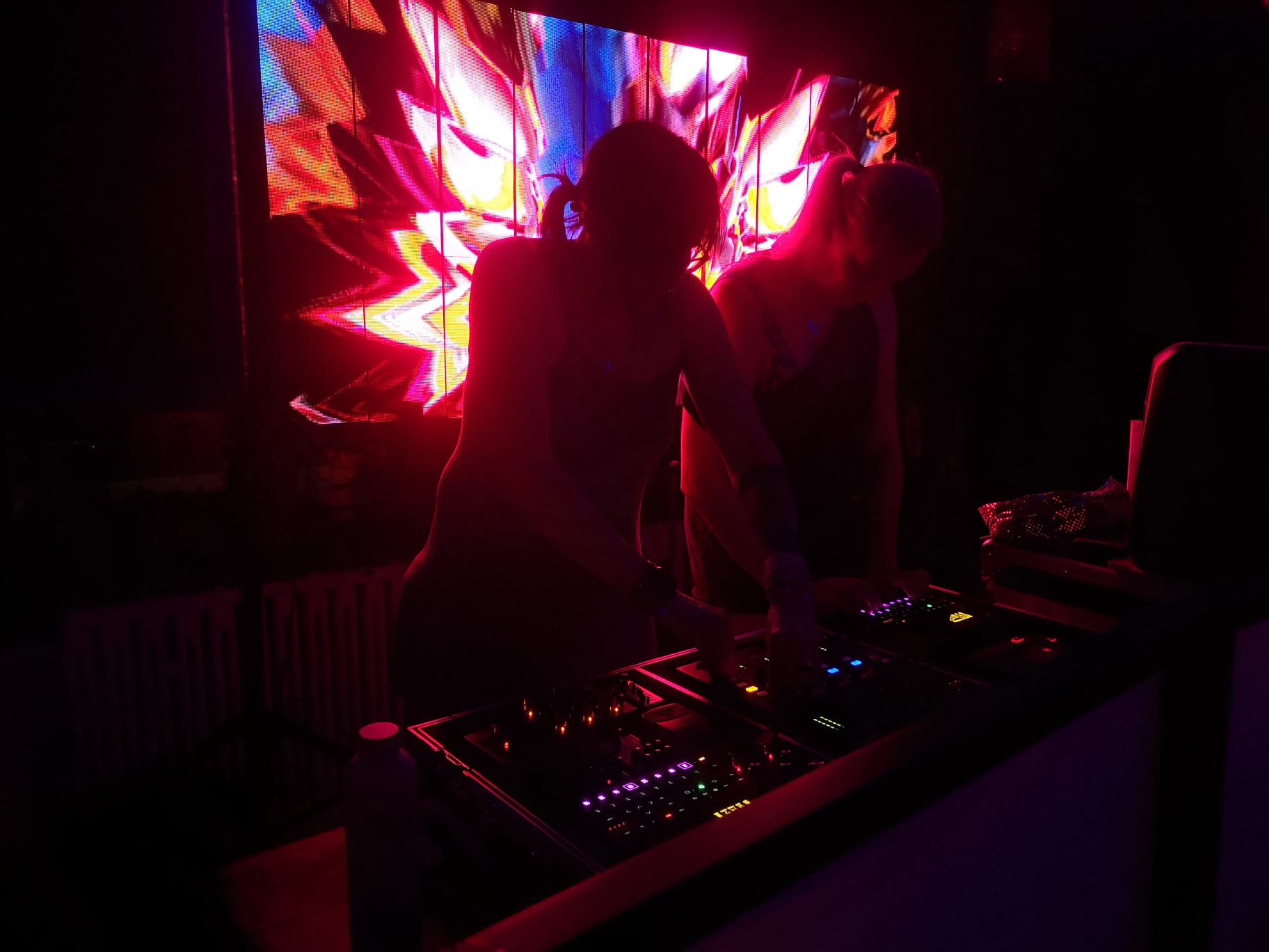Two electronic musicians performing live with synthesizers and drum machines in front of a brightly lit digital display