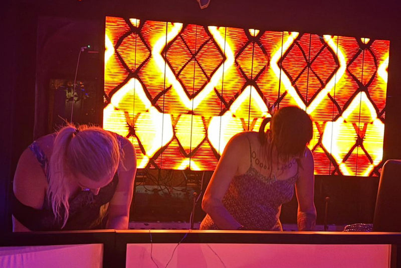 Two female electronic musicians performing live in front of an LED screen with abstract visuals
