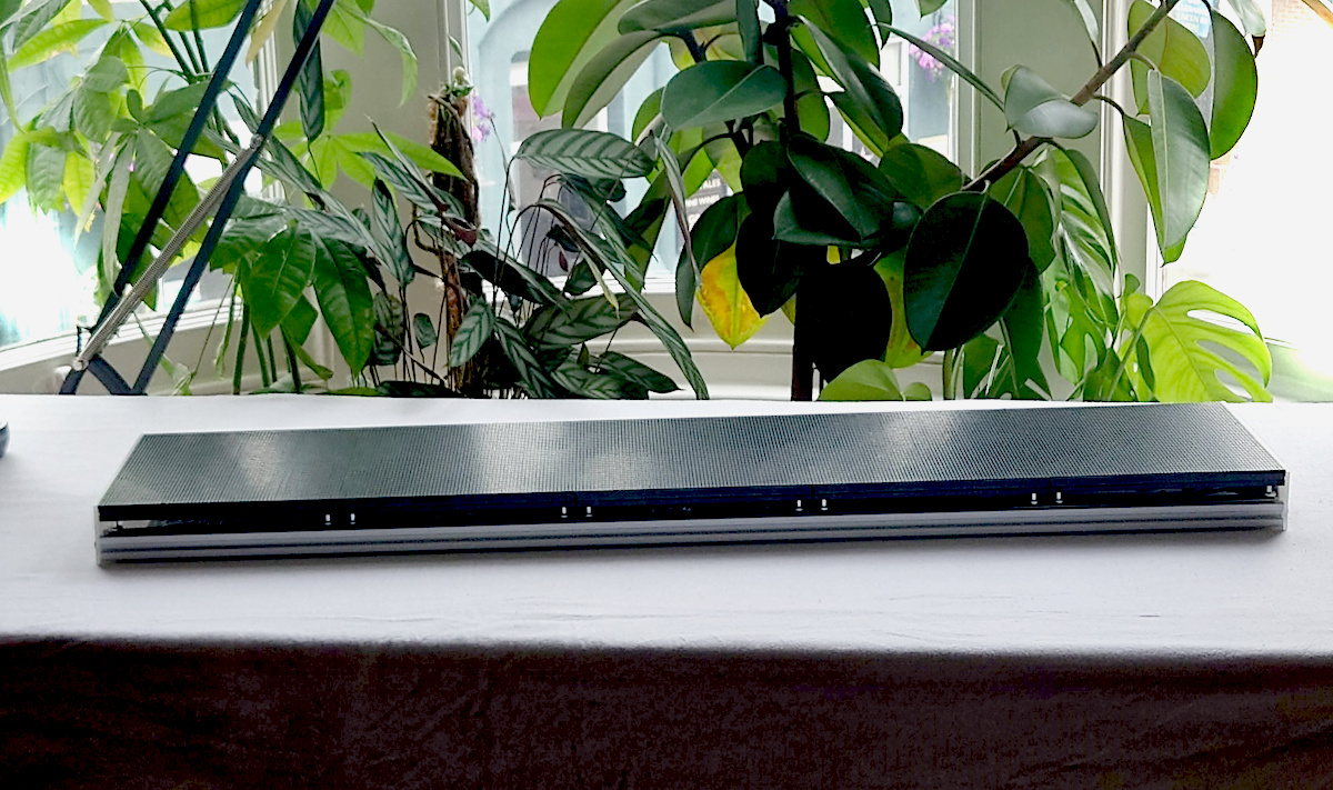 A wide shot of a long rectangular LED panel on a workbench in a bay window with houseplants in the background