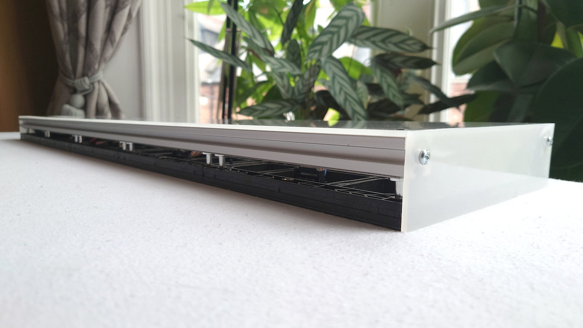 A long, narrow rectangular LED panel with white acrylic back and base, photographed on a white surface with a blurred indoor background featuring plants and curtains.