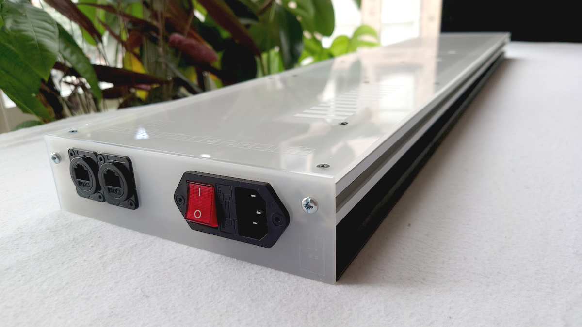A long, rectangular white LED panel with power outlets and a red power switch, photographed on a white surface with green plants in the background.