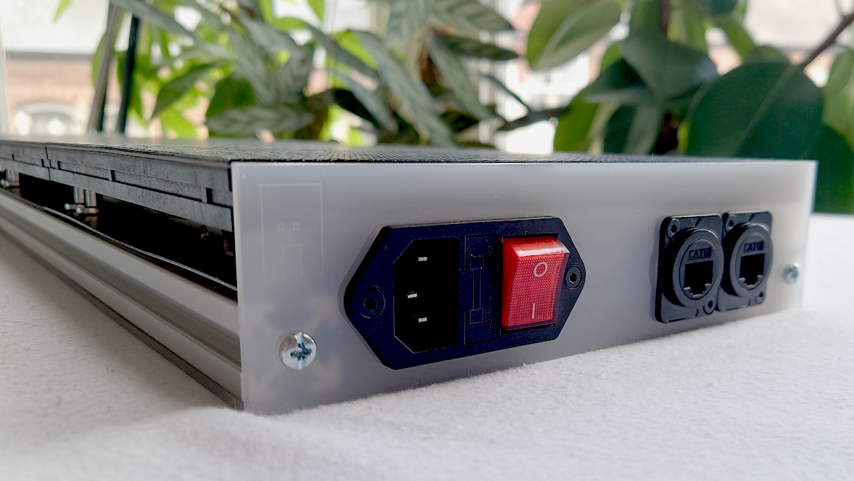 An LED panel showing the base board with a power port and 2 network ports panel mounted onto the white opaque acrylic on a white surface with green plants in the blurred background